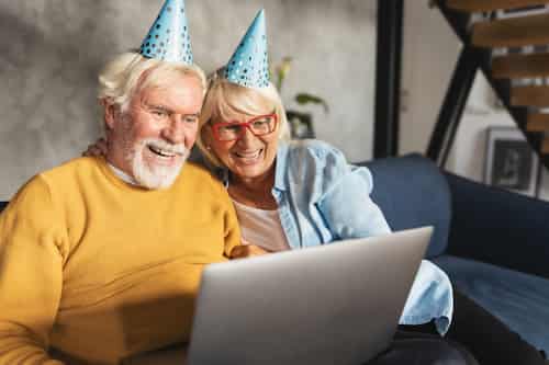 Couple enjoying virtual party with friends. Man and woman wearing party hats smiling and looking at laptop during virtual party.
